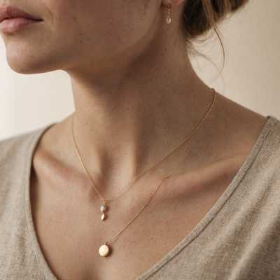 Das Bild zeigt eine Nahaufnahme des Halses und des unteren Gesichts einer Frau, die minimalistischen Schmuck trägt. Sie trägt eine zarte, vergoldete Halskette mit einem kleinen, unauffälligen Anhänger. Ihre Haut ist hell und hat einen natürlichen, makellosen Teint. Der Hintergrund ist verschwommen und neutral, wahrscheinlich in Beige- oder Hellgrautönen, wodurch der Fokus auf der Frau und ihrem Schmuck bleibt. Die Beleuchtung ist weich und gleichmäßig und hebt die Einfachheit und Eleganz des Schmucks ohne harte Schatten hervor.